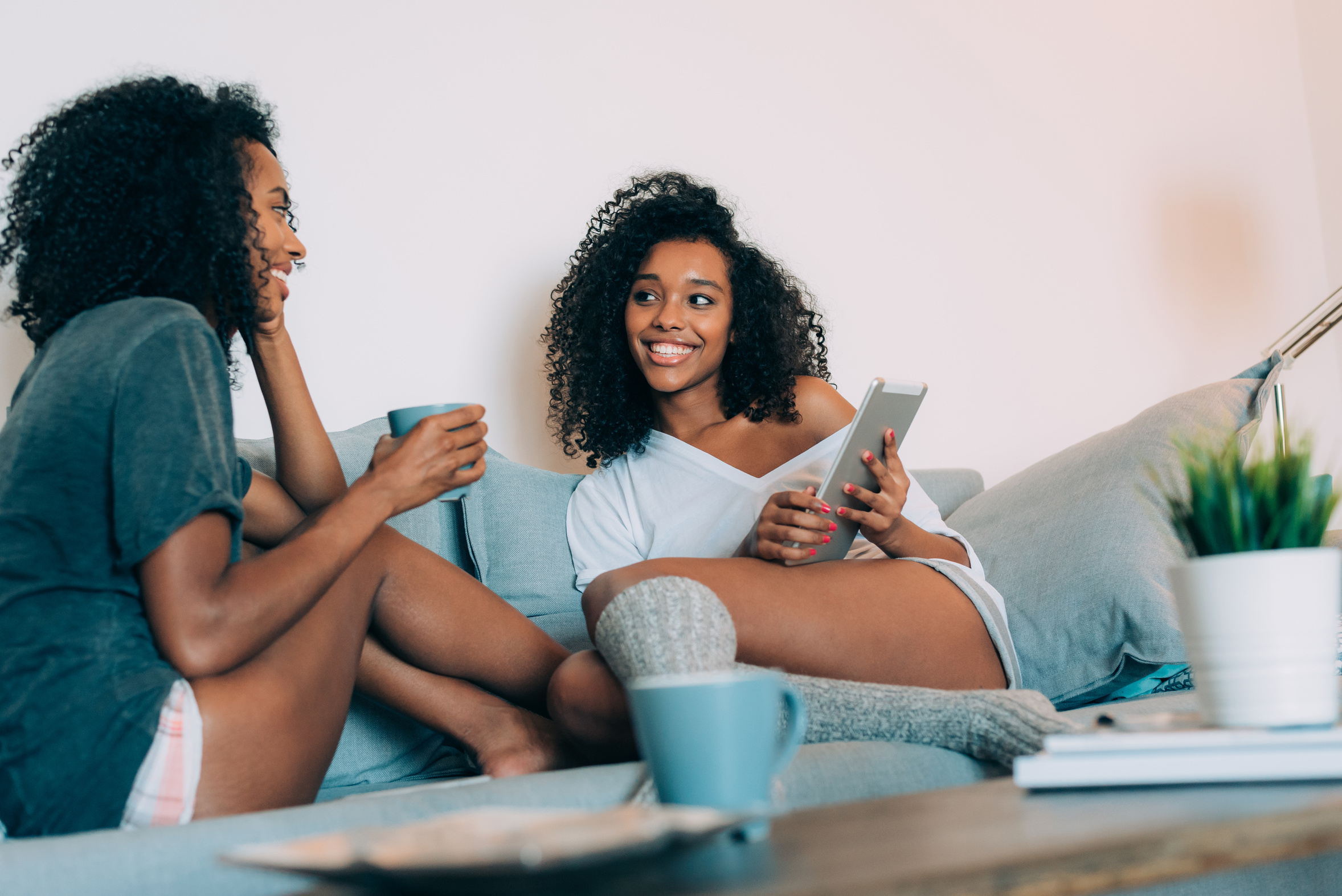 Happy young two black women sitting in the couch looking at a tablet and drinking coffee